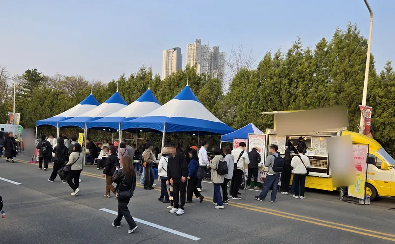 파란 천막이 늘어선 축제 거리와 노란 커피 트럭 앞에 줄 선 사람들